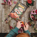 Person crafting a flower wreath on a wooden table with 'wild imagination' book and flowers.
