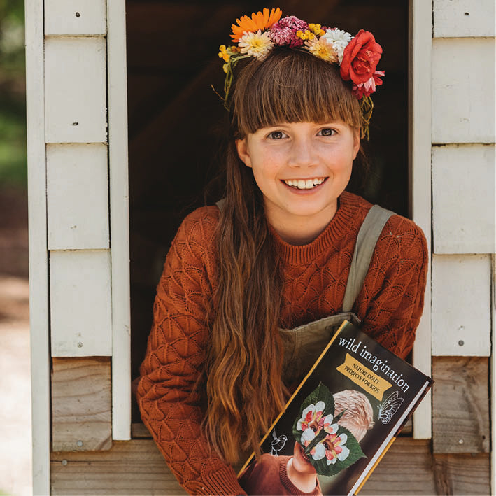 Young girl with a flower crown holding a book titled 'wild imagination' in front of a wooden door.