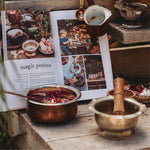Copper pots with colorful contents on a wooden surface next to an open book titled 'magic potions'.