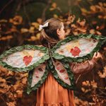 A little girl wearing leaf-like wings in a forest setting.